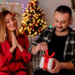 young-beautiful-couple-sitting-couch-man-opening-gift-while-his-happy-girlfriend-looking-him-celebrating-christmas-together-decorated-room-with-christmas-tree-background_141793-43564